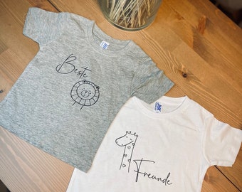May include: Two white t-shirts with black animal illustrations and text. The grey shirt has a lion with the word "Beste" above it. The white shirt has a giraffe with the word "Freunde" below it.