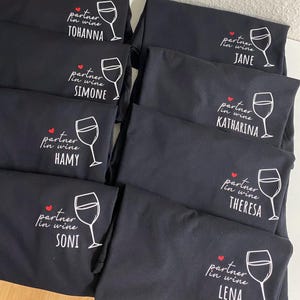 May include: Six black t-shirts with white text and a red heart. Each shirt says "partner in wine" and has a different name below:  "TOHANNA", "SIMONE", "JANE", "KATHARINA", "HAMY", "THERESA", "SONI", and "LENA".
