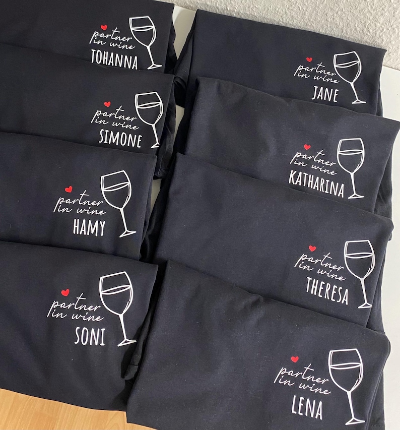 May include: Six black t-shirts with white text and a red heart. Each shirt says "partner in wine" and has a different name below:  "TOHANNA", "SIMONE", "JANE", "KATHARINA", "HAMY", "THERESA", "SONI", and "LENA".