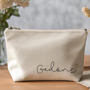 Könnte beinhalten: Cremefarbene Kosmetiktasche mit Reißverschluss und dem Wort "Gedöns" in handschriftlicher Schrift. Ein kleines Herz befindet sich neben dem Wort. Die Tasche liegt auf einer Holzoberfläche.