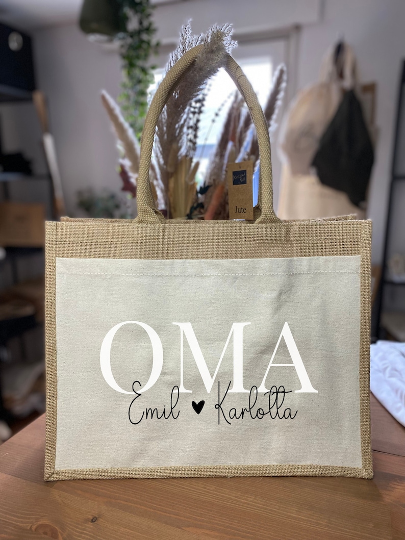 May include: A natural jute tote bag with a white canvas insert. The bag has a white handle and the text "OMA Emil &hearts; Karlotta" printed on the front.