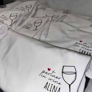 May include: A stack of white cotton tea towels with a black and red design. The design features a wine glass and the text "partner in wine" followed by a name. The names on the towels include "Sarah", "Alina", "Sven", "Gerdi", "Best", and "Stani".