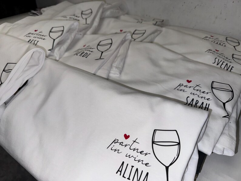 May include: A stack of white cotton tea towels with a black and red design. The design features a wine glass and the text "partner in wine" followed by a name. The names on the towels include "Sarah", "Alina", "Sven", "Gerdi", "Best", and "Stani".