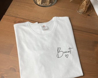 May include: White t-shirt with the word "Braut" written in black cursive with a small heart underneath.