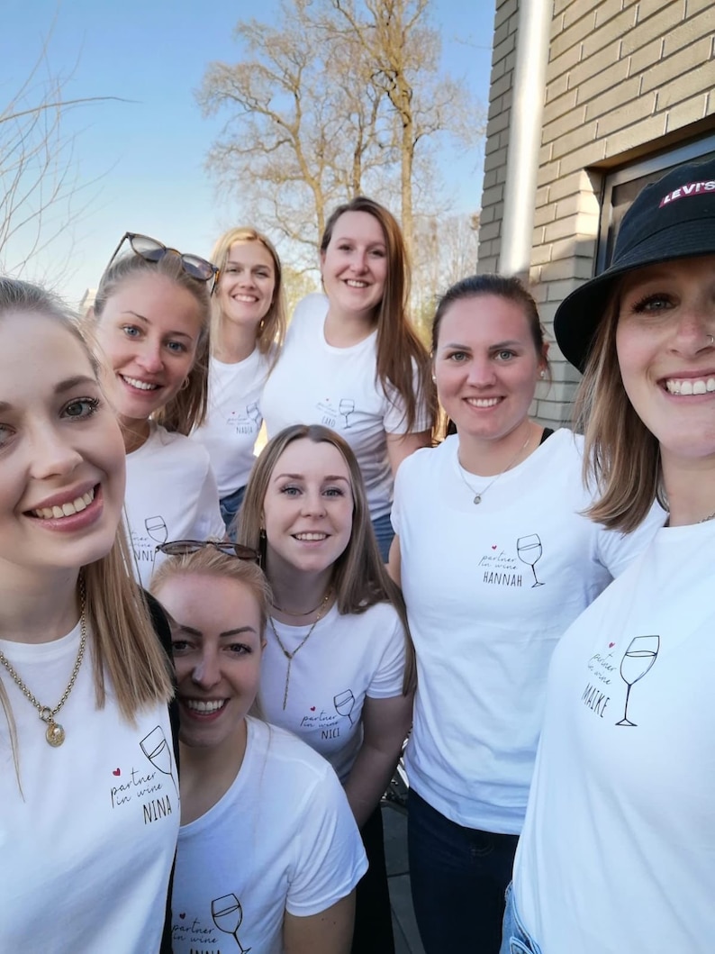 May include: A group of women wearing white t-shirts with the text "partner in wine" and their names. The women are smiling and looking at the camera.