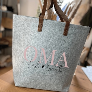 May include: A grey felt tote bag with brown leather handles. The bag has the word "OMA" printed in pink letters with the text "Emil &hearts; Karlotta" in black script below.