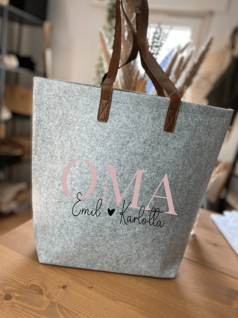 May include: A grey felt tote bag with brown leather handles. The bag has the word "OMA" printed in pink letters with the text "Emil &hearts; Karlotta" in black script below.
