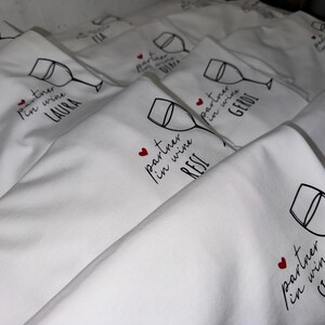 May include: A stack of white fabric tote bags with a black wine glass and the text "partner in wine" followed by a name. The text is in black with a red heart.