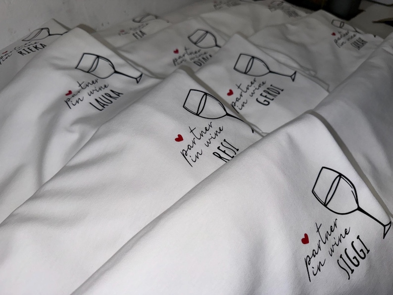 May include: A stack of white fabric tote bags with a black wine glass and the text "partner in wine" followed by a name. The text is in black with a red heart.