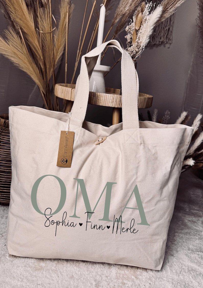 May include: A beige canvas tote bag with the word "OMA" in green letters and the names "Sophia, Finn, Merle" written below in black. The bag has a brown tag with the text "Earthwise" on it.