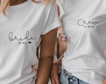 Könnte beinhalten: Zwei weiße T-Shirts mit schwarzem Text. Das erste Shirt sagt "bride to be" mit einem kleinen Herzen. Das zweite Shirt sagt "crew to drink" mit zwei Champagnergläsern.