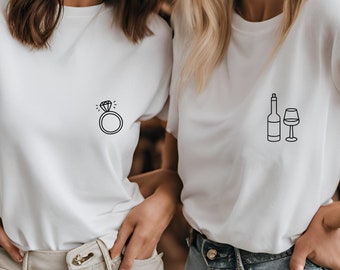 May include: Two women wearing white t-shirts with different designs. The woman on the left has a ring with a diamond on her shirt. The woman on the right has a wine bottle and glass on her shirt.