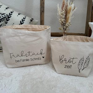 Könnte beinhalten: Zwei weiße Stoffaufbewahrungskörbe mit schwarzem Text. Der Korb links sagt "Frühstück bei Familie Schmitz" mit einem Herzsymbol. Der Korb rechts sagt "brot zeit" mit einer Weizenähre-Grafik.