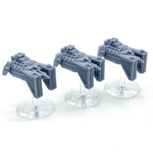 May include: Three grey miniature spaceship models, each mounted on a clear plastic stand. The spaceships have detailed, angular designs with multiple rectangular sections and a central body. The models are displayed against a white background.