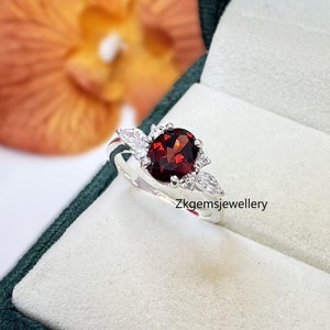 May include: A silver ring featuring a red gemstone surrounded by smaller white gemstones. The ring is displayed in a green velvet box.