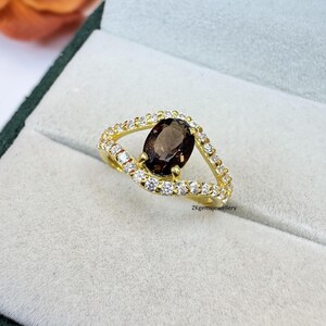 May include: A gold ring featuring a dark brown oval gemstone at the center, surrounded by a wave-like design encrusted with small, clear gemstones. The ring is set against a light gray background.