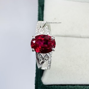 Peut inclure: Une bague en argent ornée d'une grosse pierre rouge foncé de forme ovale. La monture est ornée de plus petites pierres précieuses transparentes et de filigranes décoratifs. Le texte "ZKGEMSJEWELLERY" est visible sur la bague.