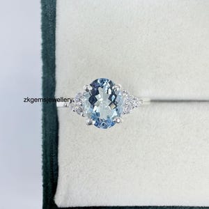 May include: A silver ring with an oval, pale blue gemstone and smaller, clear stones. The ring is displayed in a dark green jewellery box with a white interior. The text "zkgemsjewellery" is visible.