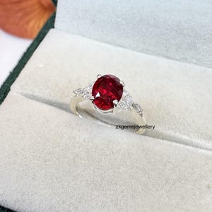 Könnte beinhalten: Ein silberner Ring mit einem großen, oval geschliffenen, tiefroten Edelstein als Mittelpunkt. Der Ring wird von kleineren, klaren Edelsteinen auf jeder Seite flankiert. Der Ring wird in einer grünen Samt-Schmuckschatulle präsentiert.