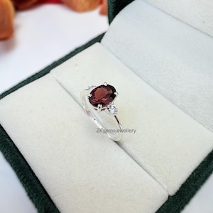 May include: A silver ring featuring a large oval red gemstone, flanked by two smaller white gemstones. The ring is displayed in a green velvet ring box.