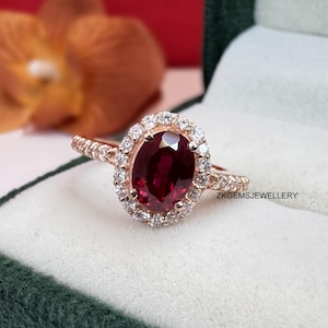May include: Rose gold ring featuring a large oval-cut, deep red gemstone surrounded by a halo of smaller, clear stones. The ring's band is also adorned with small, sparkling accents. The ring is set against a green and white background.