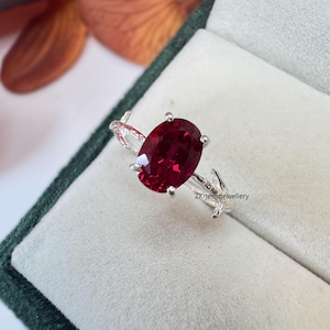 May include: A silver ring featuring a large, oval-cut, deep red gemstone held by four prongs. The band has a textured, branch-like design with small leaf details. The text ZKgemsjewellery is visible on the band near the stone setting.