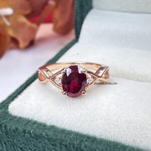 May include: A rose gold ring with a large oval, deep red gemstone as the centerpiece. The ring features a twisted band design and small, clear accent stones on either side of the main stone. The ring is displayed in a green velvet ring box.