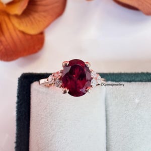 Peut inclure: Bague en or rose ornée d'une grande pierre précieuse rouge foncé de forme ovale. La bague est ornée de petites pierres précieuses claires de chaque côté de la pierre principale. La bague est posée sur un fond blanc et un écrin en velours vert foncé.