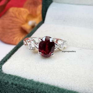 May include: A silver ring featuring a large, oval-cut, deep red gemstone. The band has small, clear accent stones and decorative filigree. The ring is presented in a green velvet box, with the text "zkgemsjewellery".