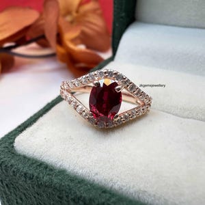 Peut inclure: Bague en or rose ornée d'un grand rubis ovale. Le rubis est entouré d'un halo de petites pierres précieuses claires. La monture de la bague a un design ouvert unique avec des pierres supplémentaires. La bague est présentée dans un écrin en velours vert.