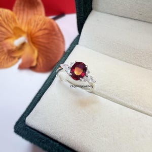 May include: A silver ring featuring a large oval red gemstone surrounded by smaller white gemstones. The ring is displayed in a green velvet ring box. ZKgemsjewellery