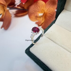 May include: A silver ring featuring a large oval red gemstone, surrounded by smaller white gemstones. The ring is set in a delicate floral design.