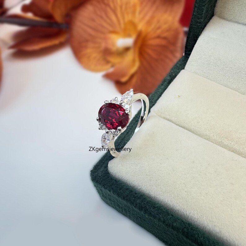 May include: Close-up of a sterling silver ring featuring an oval-shaped, dark red gemstone, possibly a ruby or garnet, encircled by smaller, clear cubic zirconia stones.  The ring's band is thin and delicate. The brand name 'ZKgemsjewellery' is subtly visible. The ring rests in a dark green jewelry box, providing a sophisticated presentation. The setting is elegant and the overall style is classic and refined.