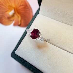 May include: A silver ring featuring a large oval ruby gemstone, surrounded by smaller white gemstones. The ring is displayed in a white velvet ring box. The text "ZKgemsjewellery" is printed on the box.