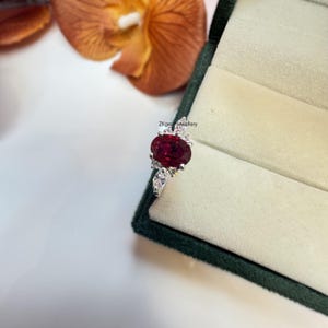 May include: A silver ring featuring a large oval ruby gemstone, surrounded by smaller white gemstones. The ring is presented in a green velvet ring box.