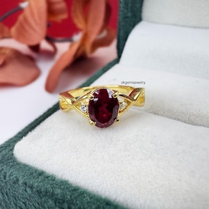 Könnte beinhalten: Ein Goldring mit einem zentralen, oval geschliffenen, tiefroten Edelstein in einer Vier-Krappen-Fassung, flankiert von zwei kleinen klaren Steinen auf jeder Seite. Die Ringschiene hat ein verschlungenes Design. Der Text zkgemsjewelry ist sichtbar.