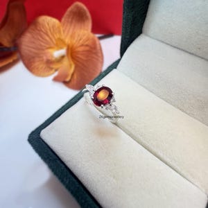 May include: A silver ring featuring a red oval gemstone surrounded by smaller white gemstones. The ring is set in a leaf design. The ring is displayed in a green velvet box. ZKgemsjewellery.