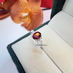 May include: A silver ring featuring a red oval gemstone surrounded by smaller white gemstones. The ring is set in a leaf-like design. The ring is displayed in a green velvet box. ZKgemsjewellery.