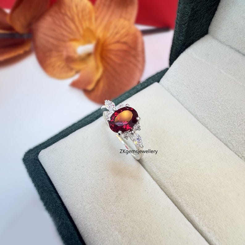 May include: A silver ring featuring a red oval gemstone surrounded by smaller white gemstones. The ring is set in a leaf-like design. The ring is displayed in a green velvet box. ZKgemsjewellery.