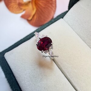 May include: A silver ring featuring a large oval ruby gemstone surrounded by smaller white gemstones. The ring is displayed in a green velvet ring box. The text "ZKgemsjewellery" is visible on the box.