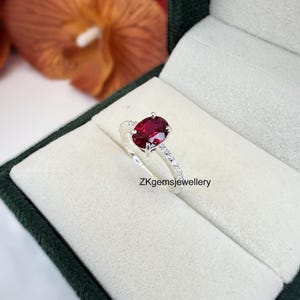 May include: A silver ring with a large oval-shaped red gemstone surrounded by smaller white gemstones. The ring is in a white box with a green lining. ZKgemsjewellery is printed on the box.