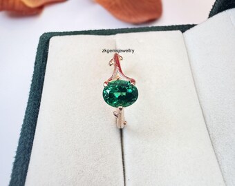May include: A rose gold ring featuring a large, oval-cut emerald gemstone. The ring has a delicate, ornate design with a decorative top. The ring is set against a white background inside a dark green jewelry box.