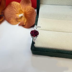 May include: A silver ring featuring a large oval red gemstone, surrounded by smaller white gemstones. The ring is displayed in a green velvet ring box.