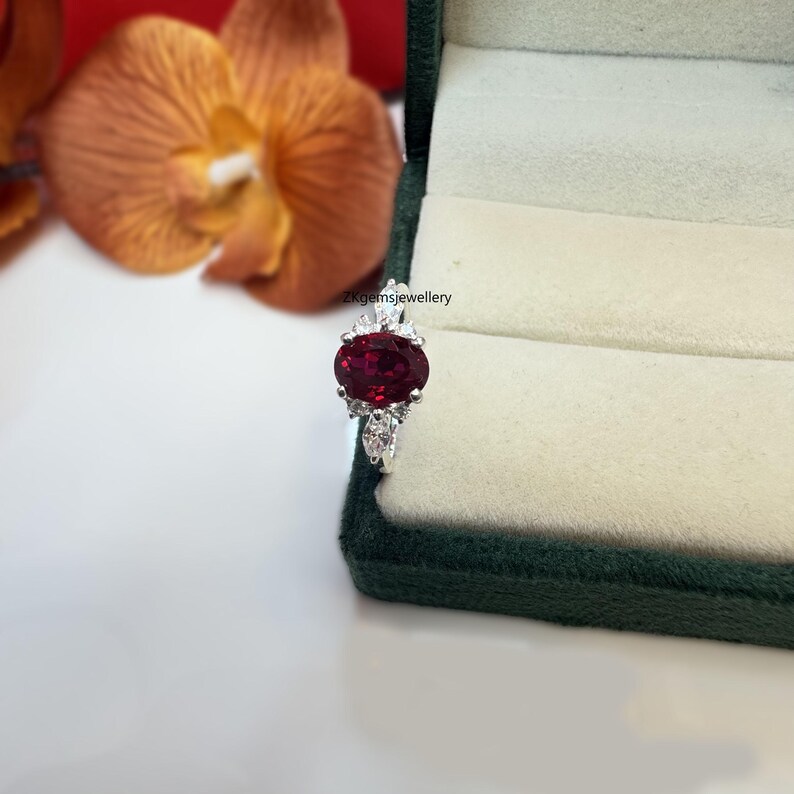 May include: A silver ring featuring a large oval red gemstone, surrounded by smaller white gemstones. The ring is displayed in a green velvet ring box.