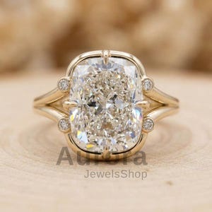 May include: A gold ring featuring a large, rectangular, clear gemstone. The ring has a split shank design and small accent stones. The text "Aurora JewelsShop" is visible at the bottom of the image.