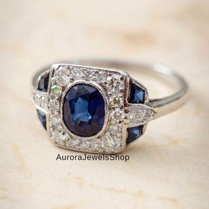 Op de afbeelding: Een zilveren ring met een ovale saffier in het midden, omringd door kleine diamanten en vierkante saffieren. De ring heeft een vintage design, met de tekst "AuroraJewelsShop".