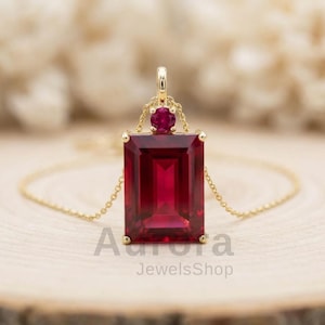 May include: A gold-colored necklace with a rectangular, deep red gemstone pendant. A smaller, round red gemstone sits above the larger one. The pendant is suspended from a delicate gold chain. The text "Aurora JewelsShop" is visible at the bottom.