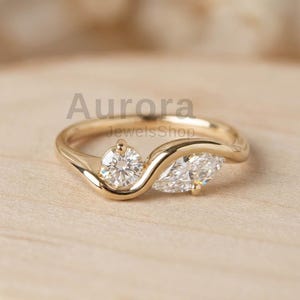 Peut inclure: Une bague en or avec deux pierres précieuses claires. L'une est ronde, l'autre en forme de marquise. L'anneau de la bague épouse les pierres. Le texte "Aurora JewelsShop" est visible.