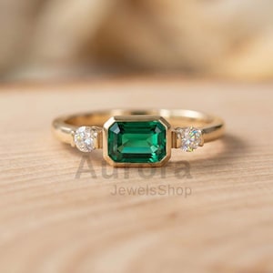 May include: A gold ring featuring a rectangular emerald-cut green gemstone in the center, flanked by two smaller, round, clear gemstones. The ring is set on a wooden surface. The text "Aurora JewelsShop" is visible.