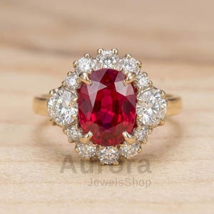 May include: A gold ring featuring a large, oval-cut ruby center stone surrounded by a halo of smaller diamonds. Two larger diamonds flank the ruby. The ring is set on a light-colored wooden surface.